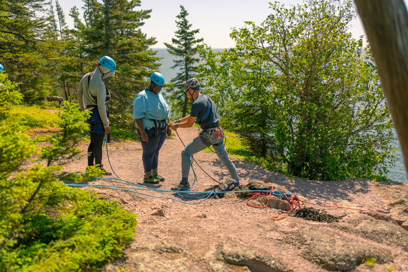 The image shows three people preparing for rock climbing or rappelling on a rocky outcrop. They are wearing helmets and harnesses, and one person appears to be adjusting another's gear. Ropes and other climbing equipment are visible on the ground. The setting is outdoors with trees and a body of water in the background.